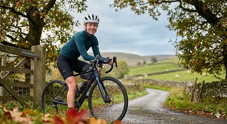 A British female cyclist in a lightweight road cycling helmet and teal jersey smiles while paused on a Peak District country lane, leaning on her road bike by a rustic wooden gate.