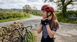 A British female cyclist adjusting the chin strap of her matte red road cycling helmet, with her long ponytail positioned comfortably at the back.